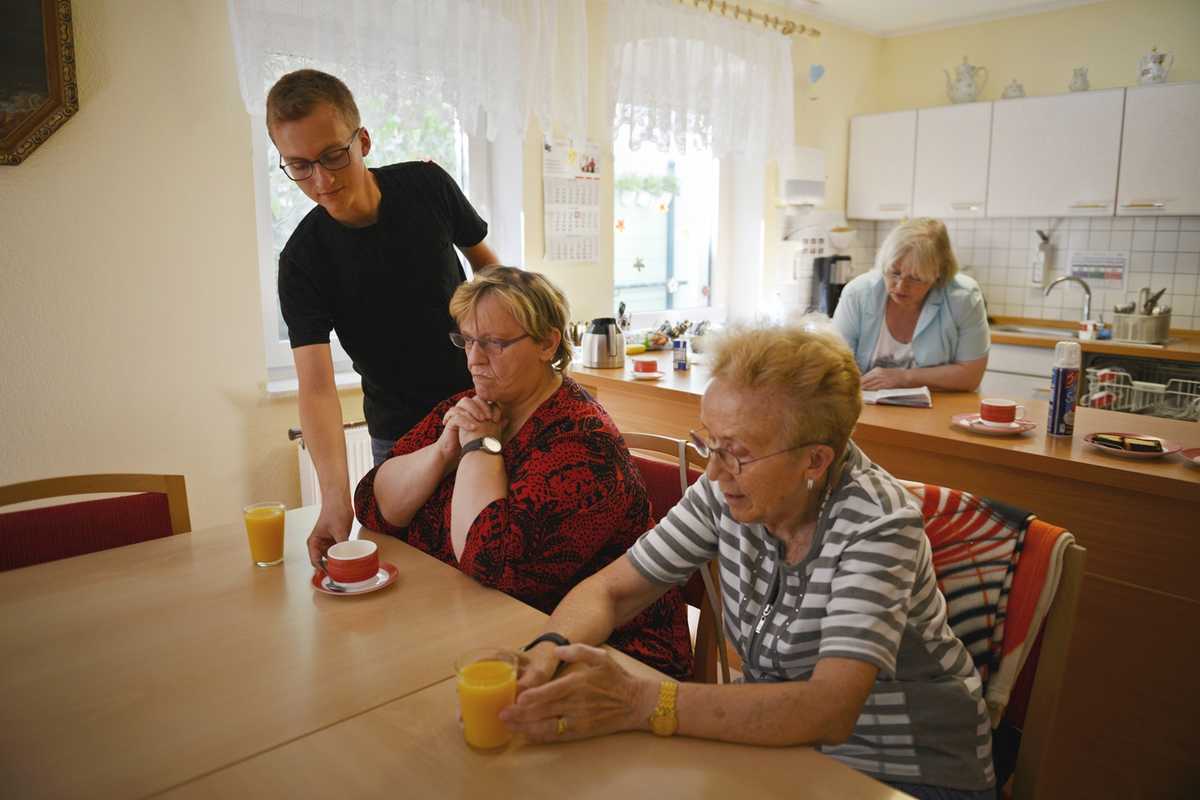 FSJ'ler Clemens Brambach bringt zwei älteren Frauen, die an einem Tisch sitzen, eine Tasse Tee.