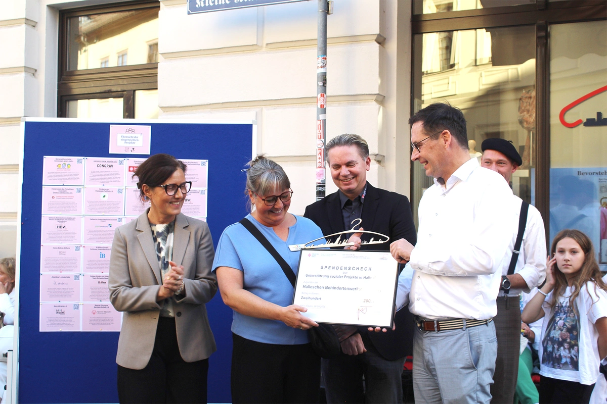 Ein Mann mit Brille und weißem Hemd und Jeans überreicht einer Frau mit Brille und blauem T-Shirt einen Spendenscheck. Daneben stehen eine Frau in einem hellen Blazer und mit Brille  und ein Mann in einem schwarzen Anzug, die beide fröhlich applaudieren.