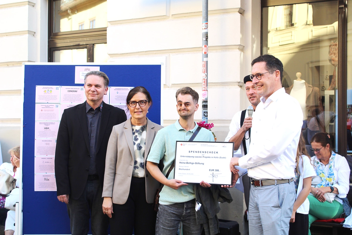 Ein Mann mit Brille und weißem Hemd überreicht einem jungen Mann in mintgrünen T-Shirt einen Spendenscheck. Links daneben steht ein Mann in schwarzem Anzug und eine Frau mit hellem Blazer und Brille. Alle gucken freundlich in eine Kamera.
