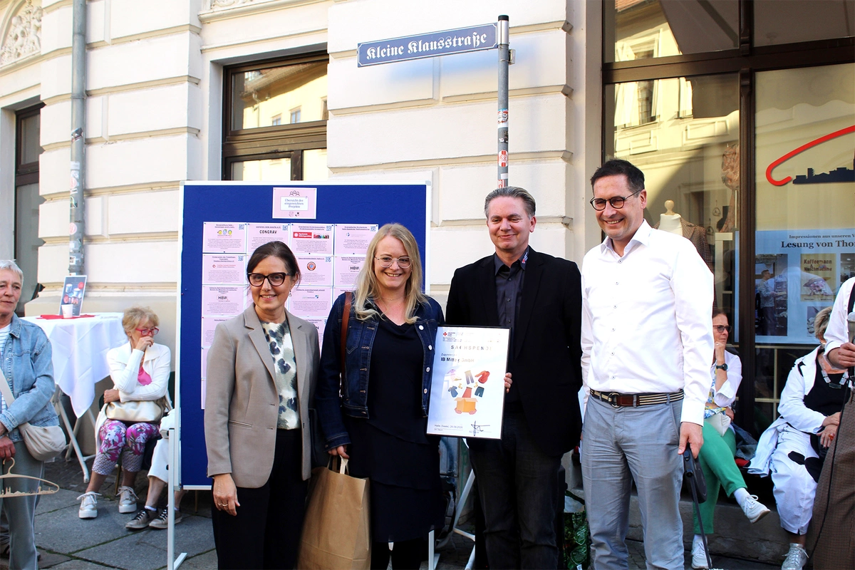 Eine Frau mit Brille und dunklen Haaren, eine Frau mit Brille und offenen blonden Haaren, ein Mann in schwarzem Anzug und ein Mann mit Brille und weißem Hemd posieren für ein Bild. Der Mann im schwarzen Anzug hält ein Spendenscheck für eine Sachspende in Form von Kleiderschenkungen in der Hand.