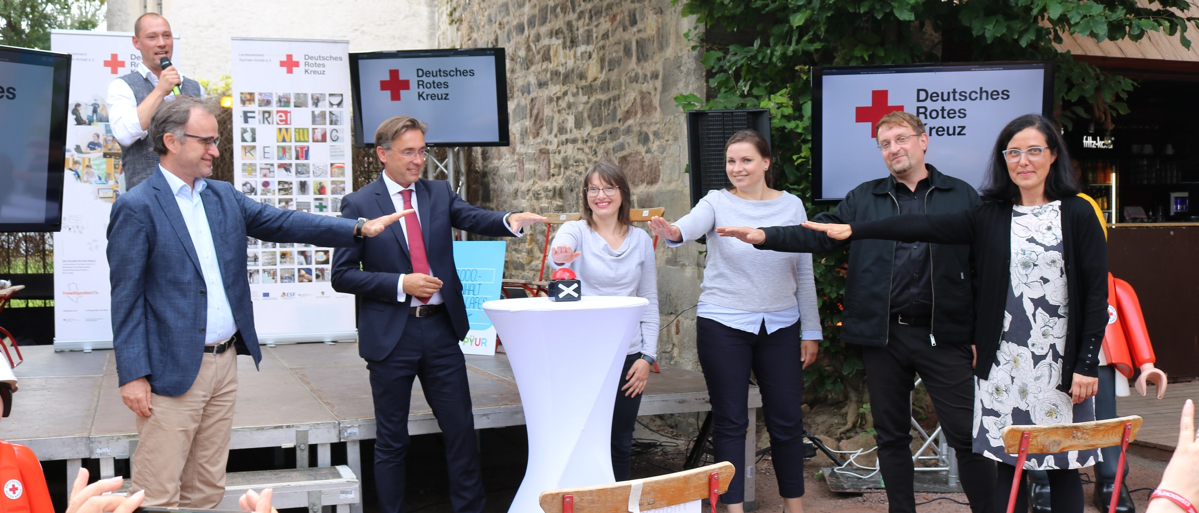 Menschen stehen vor einem Buzzer und deuten an darauf zu drücken.