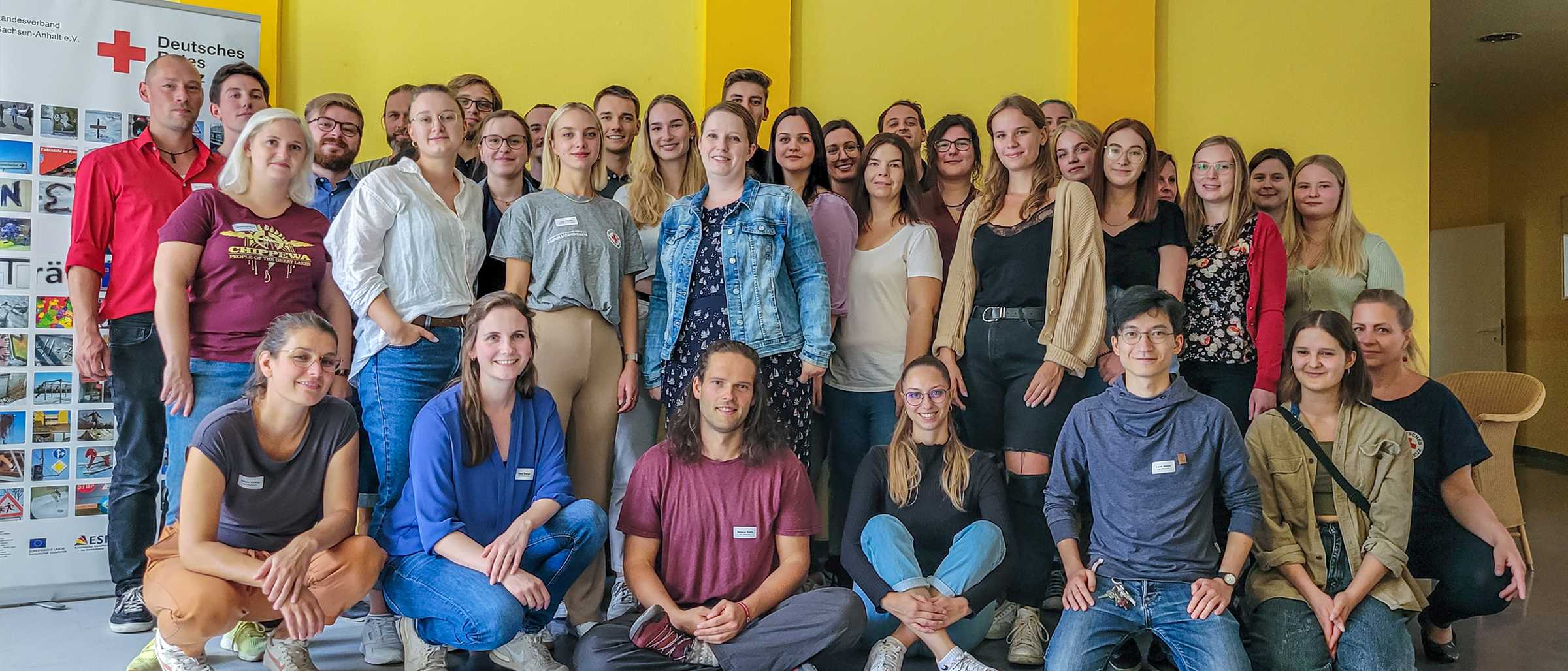 Die 22 angehenden Teamer:innen und die Mitarbeitenden des Betriebsteils Freiwilligendienste stehen und knien für ein Gruppenfoto vor einer gelben Wand in unserer Seminarakademie