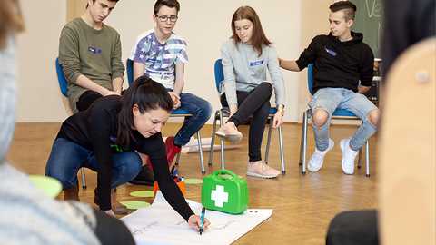 Verrückt, na und? - Schultag Foto in einem Klassenraum. Zu sehen sind vier Jugendliche in einem Stuhlkreis im Hintergrund. Im Vordergrund ist eine Schüler:in, die auf dem Boden auf ein Flipchart Papier schreibt. Neben ihr steht ein hellgrüner Erstehilfe-Kasten